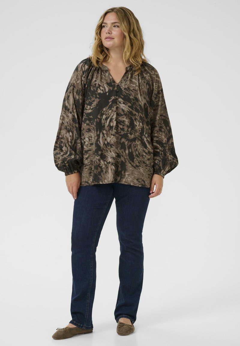 Donna con capelli biondi ondulati, che indossa una blusa marrone a fantasia larga, jeans blu scuro e ballerine marroni, in piedi su uno sfondo bianco.
