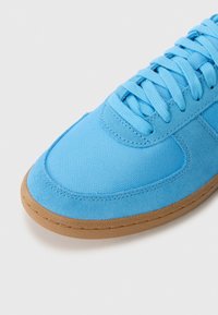 Lysblå sneakers lavet af tekstureret stof og ruskind, med flade snørebånd, en rund tå og en brun gummisål.