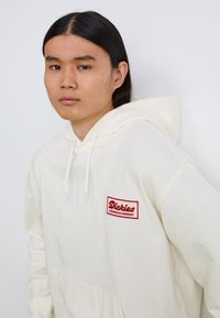 Kremfarget hettegenser med frontlomme, snor i hetten og rød brodert "Dickies Workwear Company"-lapp på venstre bryst. Glatt tekstur.