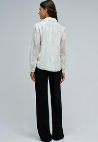 Camicia bianca e trasparente con motivo floreale testurizzato e maniche lunghe, abbinata a pantaloni larghi neri con dettaglio dei polsini a bottone.