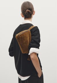 Brown suede shoulder bag with a rectangular shape and zip closure, worn over a black sweater, showcasing a minimalist design.