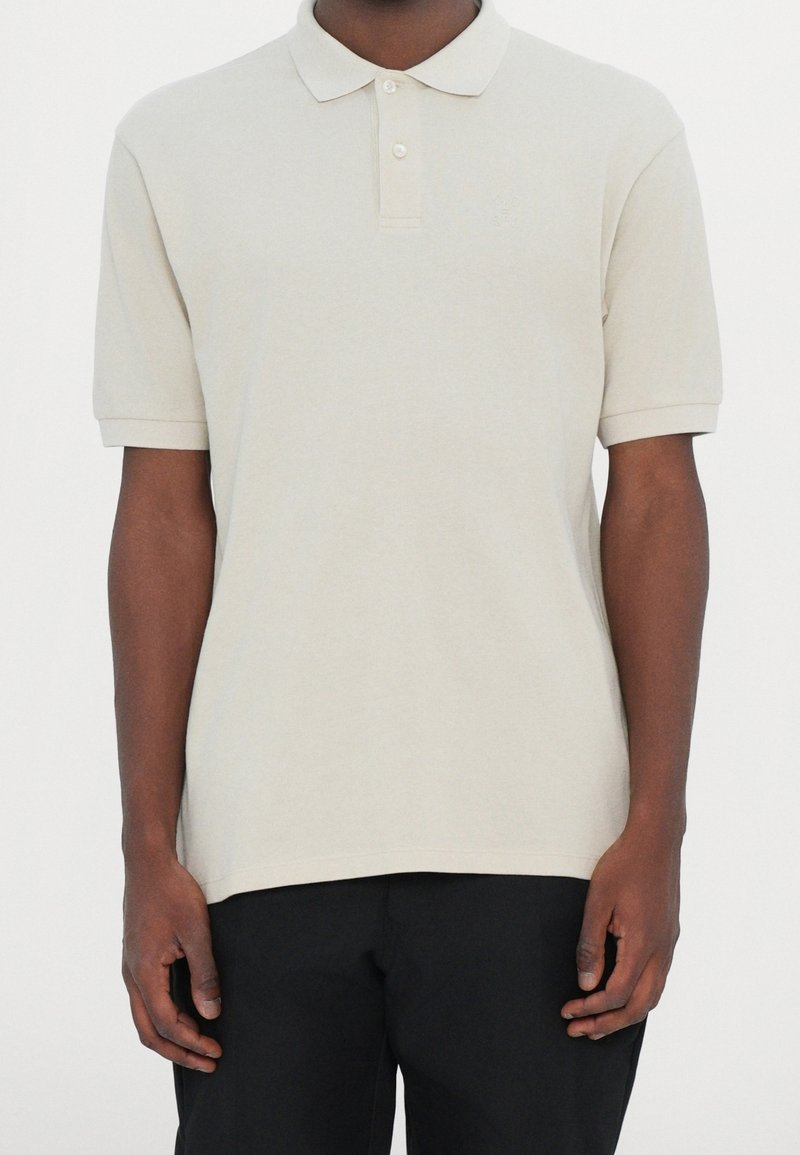 Homme portant un polo beige clair à manches courtes avec deux boutons, assorti à un pantalon noir, montré du cou jusqu'aux hanches.
