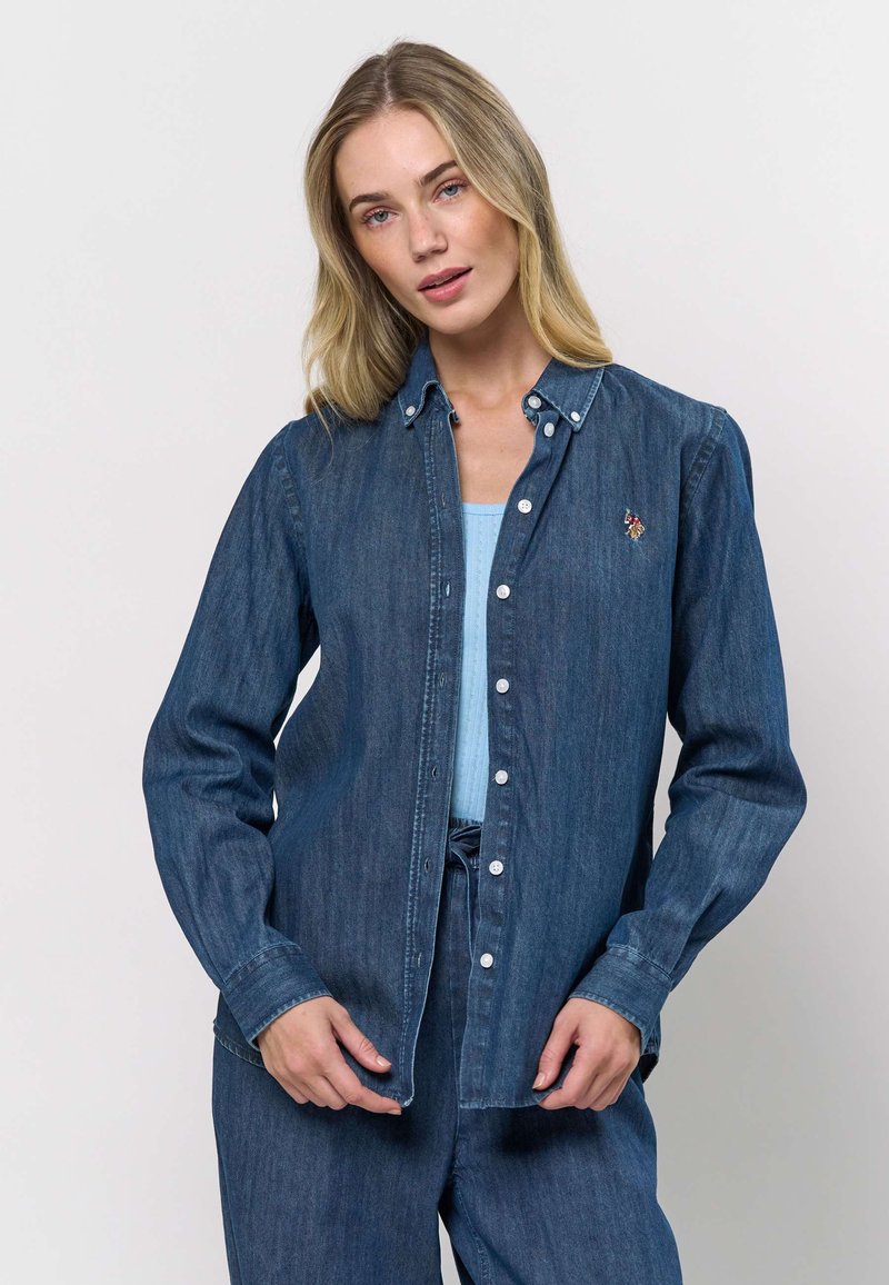 Cămașă din denim cu nasturi, de culoare albastru închis, cu guler clasic, mâneci lungi și un detaliu floral brodat pe piept.