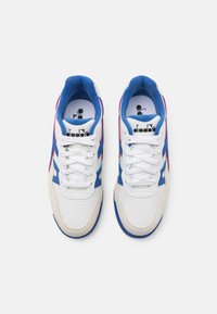 White and blue sneakers featuring a textured upper, perforated material, and colorful accents in red and blue. Laces and branding visible.