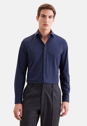 Jeune homme portant une chemise bleu marine boutonnée et un pantalon habillé gris foncé, debout devant un fond clair uni.