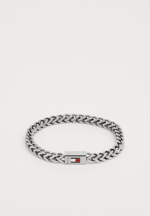 Bracelet en chaîne argentée avec des maillons entrelacés et un fermoir rectangulaire comportant des détails en incrustation rouge et bleue. Texture métallique lisse.