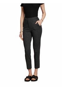 Žena v tmavě šedých slim-fit kalhotách s vysokým pasem a bočními kapsami, černém topu s krátkým rukávem, zlatých náramcích a černých sandálech s otevřenou špičkou.