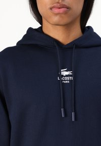 Tamsiai mėlyna džemperio tipo palaidinė iš medvilnės mišinio su raišteliu. Papuošta baltu "LACOSTE PARIS" logotipu ir krokodilo grafika ant krūtinės. Palaidinės paviršius lygus.