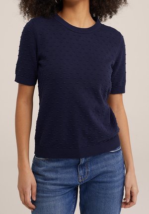 Femme portant un haut en maille texturée bleu marine à manches courtes avec un motif à pois et un jean bleu sur un fond neutre.