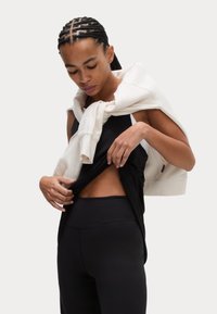 Svart atletisk tanktopp med sidoutskärningar, lager-på-lager under en krämfärgad sweatshirt som är slängd över axlarna. Svarta högmidjade leggings.