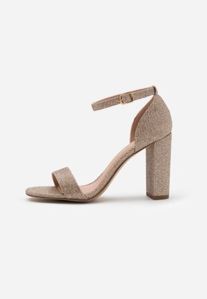 Błyszczące złote sandały na obcasie z paskiem na kostce, odkrytymi palcami i grubym obcasem. Teksturowana tkanina i gładka wkładka.