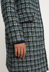 Ullblandad kappa med ett blått, svart och brunt rutmönster, struktur med en fluffig finish och en sidficka.