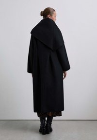 Abrigo largo negro con cuello alto, hecho de tela suave. Presenta un ajuste holgado y sobredimensionado con detalles sutiles en las costuras. Llevado sobre un vestido estampado.