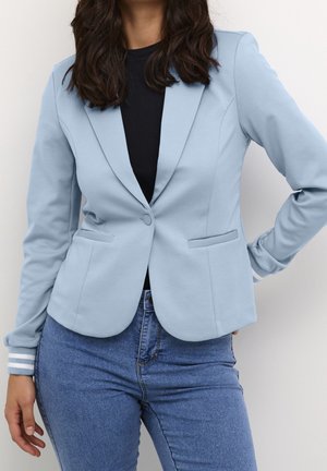 Kobieta w jasnoniebieskiej marynarce, czarnym topie i niebieskich dżinsach, stoi z jedną ręką na biodrze na jednolitym tle.