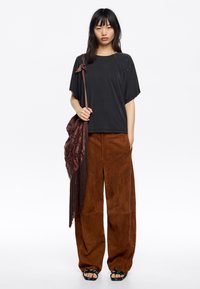 Camiseta negra de corte holgado con escote redondo y adornos en los hombros, combinada con pantalones de ante marrón de tiro alto y anchos, y sandalias negras.