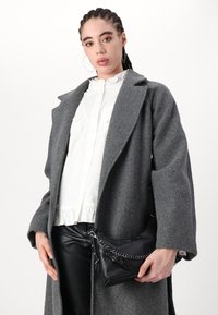 Grau melierter Wollmantel mit breiten Revers, kombiniert mit einer weißen Rüsch Bluse und schwarzen Lederhosen. Schwarze Handtasche mit Kettenakzenten.