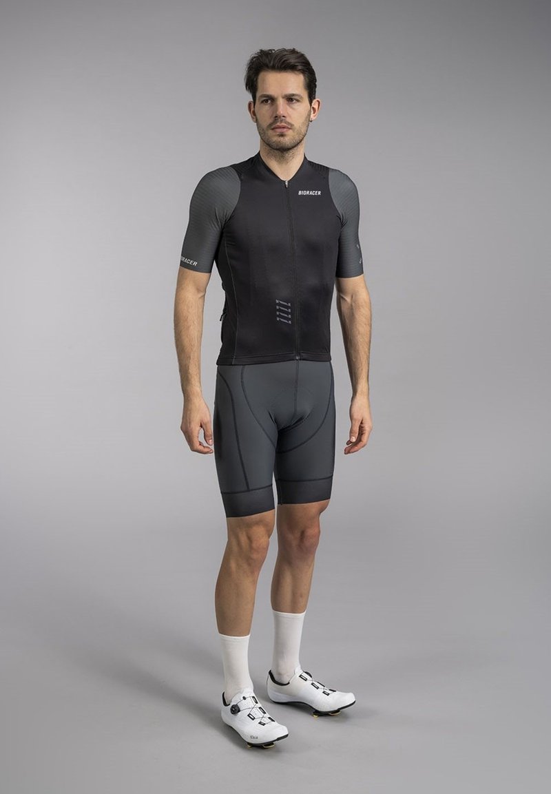 Zwart kortshirt fietsbody met grijze accenten, slim fit, ritsluiting, gecombineerd met donkere shorts en witte fietsschoenen.