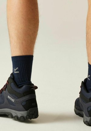 Nogi w granatowych butach turystycznych z czarnymi podeszwami i granatowymi skarpetkami stojące na jasnej neutralnej podłodze na tle jednolitej ściany.