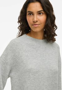 Jauna sieviete ar krokainiem tumšiem matiem, ģērbta gaiši pelēkā teksturētā džemperī, nedaudz skatās pa labi uz vienkrāsainas baltas fona.