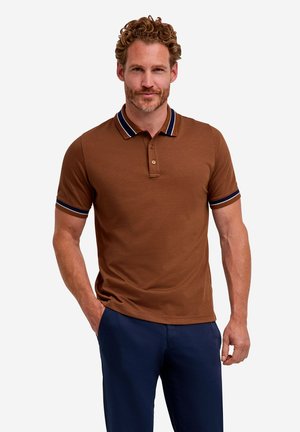 Uomo con capelli ricci che indossa una polo marrone a maniche corte con bordi blu navy e pantaloni blu navy, in piedi con una mano in tasca su sfondo bianco.