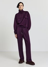 Lila, gestrickter Trainingsanzug mit Reißverschluss am Stehkragen, zwei Brusttaschen und schmal zulaufender Hose mit elastischem Bund, kombiniert mit dunklem Schuhwerk.