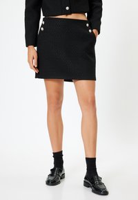 Falda mini de tela negra tejida con dos botones decorativos plateados en la parte delantera. Combinada con calcetines negros y mocasines brillantes.