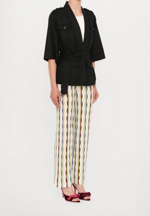 Personne portant une veste noire ceinturée, un top blanc, un pantalon large rayé et des talons ouverts bordeaux avec des accents de nœud, debout devant un fond uni.
