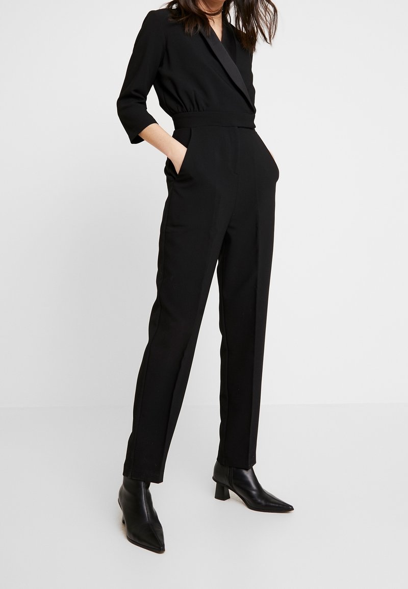 Femme portant une combinaison noire cintrée avec manches 3/4 et bottines noires pointues, debout les mains dans les poches.