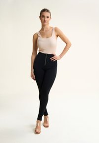 Schwarze hochtaillierte Hose mit einem Reißverschluss auf der Vorderseite, kombiniert mit einem figurbetonten beigefarbenen Tanktop. Durchsichtige Sandalen mit Absatz runden den Look ab.