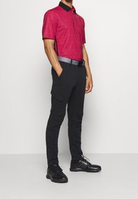 Červené polo tričko s krátkým rukávem a černým límečkem, kombinované s černými kargo kalhotami s bočními kapsami a šedým pásem, nošené s černými teniskami.