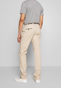 Beige chinos with a straight leg design, featuring two back pockets with zippers, and a black and green striped belt. Paired with white shoes.