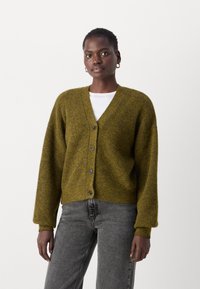 Ελαιόχρωμο cardigan φτιαγμένο από υφασμάτινη πλέξη με υφή, διαθέτει V-λαιμό, πέντε μαύρα κουμπιά και φαρδιά μανίκια, συνδυασμένο με γκρίζα τζιν.