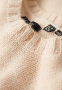 Pull en tricot rose clair avec un col à côtes. Étiquettes visibles indiquant la taille : UK 10, US 6, UE 38. Texture douce avec une finition légèrement duveteuse.