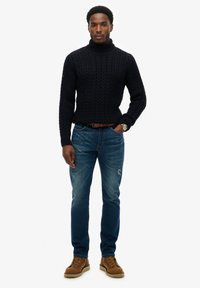 Schwarzer Kabelstrick-Turtleneck-Pullover, blaue abgenutzte Jeans und braune Schnürstiefel. Uhr am linken Handgelenk getragen. Einfaches, lässiges Outfit.