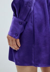 Lila Satin-Kleid mit glänzender Textur, ausgestattet mit einem Knopfdetail am Ärmel und einer glatten, fließenden A-Linie-Silhouette.
