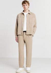 Giacca camicia beige con bottoni e due tasche sul petto, indossata sopra una camicia bianca e pantaloni beige abbinati, completata da sneakers bianche.