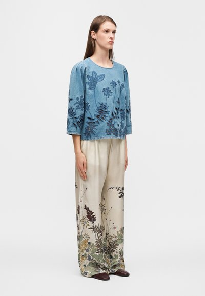 Haut en denim avec broderies florales, associé à un pantalon large de couleur claire avec motifs botaniques. Des chaussures marron complètent la tenue.