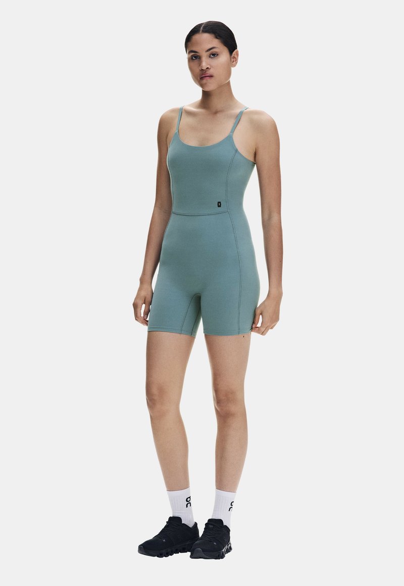 Femme debout portant une combinaison sans manches bleu sarcelle avec des bretelles fines, des chaussettes blanches et des chaussures de sport noires sur un fond clair.