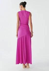 A woman stands with her back to the camera, wearing a fitted pink maxi dress that flares at the bottom, complemented by beige heels.