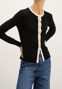 Black cardigan with cream buttons and trim, featuring a fitted design and long sleeves. The fabric appears smooth and lightweight.