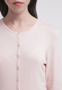 Cárdigan rosa claro con cuello redondo, cierre delantero con botones y textura de punto suave. Con seis botones en el centro.