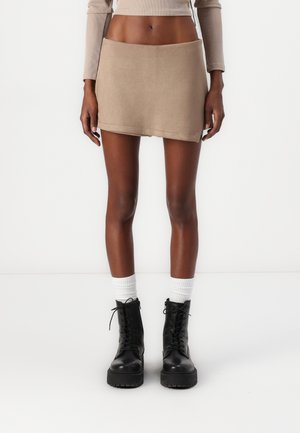 Slank person iført en beige mininederdel, cropped langærmet top, hvide sokker og sorte snørebånds kampstøvler, stående på en hvid baggrund.