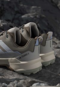 Paire de chaussures de randonnée Adidas Terrex beige et blanche sur un sol rocheux, montrant des collerettes rembourrées et des semelles robustes pour une utilisation en extérieur.