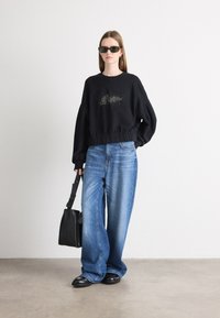 Felpa nera a maniche lunghe con dettagli metallici, jeans blu in denim larghi, borsa nera e occhiali da sole scuri. Texture morbide e vestibilità rilassata.