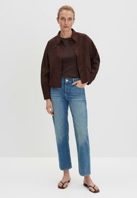 Bruine suède cropped jas met knoopsluiting, gecombineerd met lichtblauwe rechte jeans en zwarte flip-flops, tegen een effen achtergrond.
