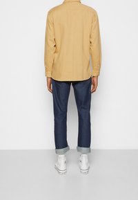 Chemise à manches longues de couleur moutarde avec col et poignets boutonnés, associée à un jean bleu marine retroussé et des baskets blanches.