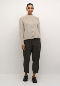 Beige, textuurgebreide cardigan met een hoge kraag, voorzien van vijf grote zilveren knopen, gedragen over donkere, taps toelopende broeken en zwarte loafers.