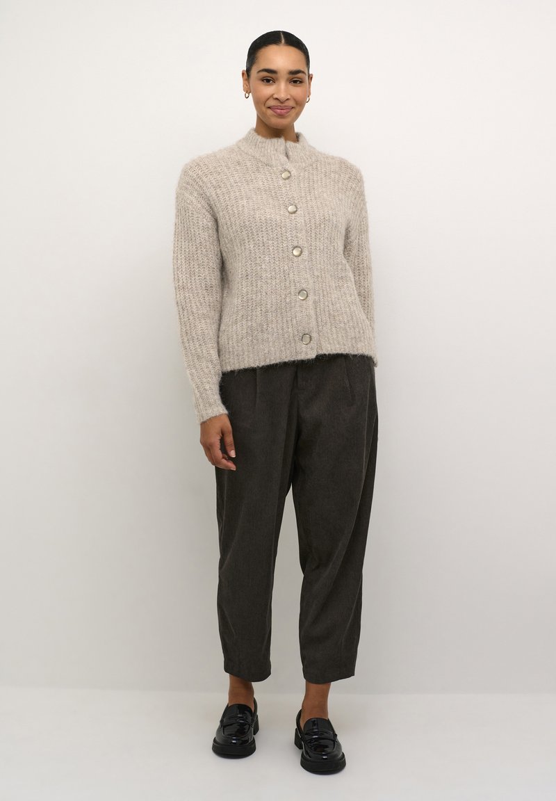 Beige, textuurgebreide cardigan met een hoge kraag, voorzien van vijf grote zilveren knopen, gedragen over donkere, taps toelopende broeken en zwarte loafers.