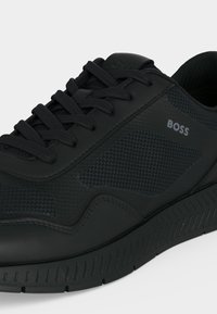 Baskets basses noires avec panneaux latéraux texturés, lacets noirs et logo « BOSS » sur le côté extérieur au-dessus de la semelle.