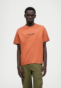 Tricou cu mânecă scurtă din bumbac portocalie ruginită, cu guler rotund. Prezintă textul alb "RIPCURL SURF COMPANY SINCE SIXTY NINE" pe față.